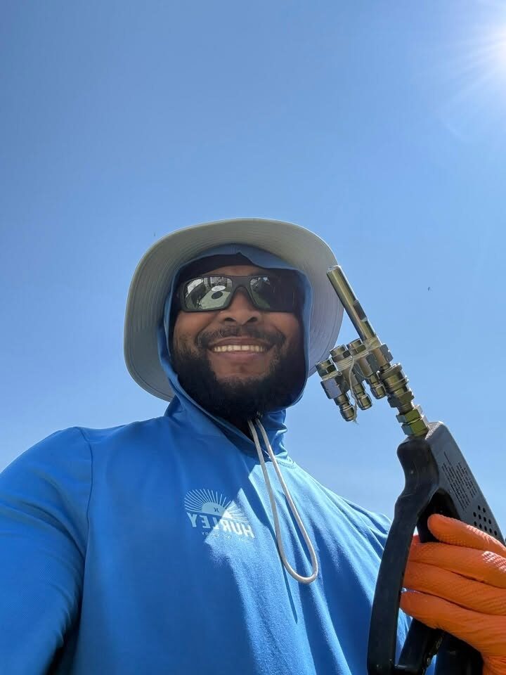 The owner of Lucky Duck Pressure Washing on a job, smiling in a sun hat with a pressure washing gun over his shoulder